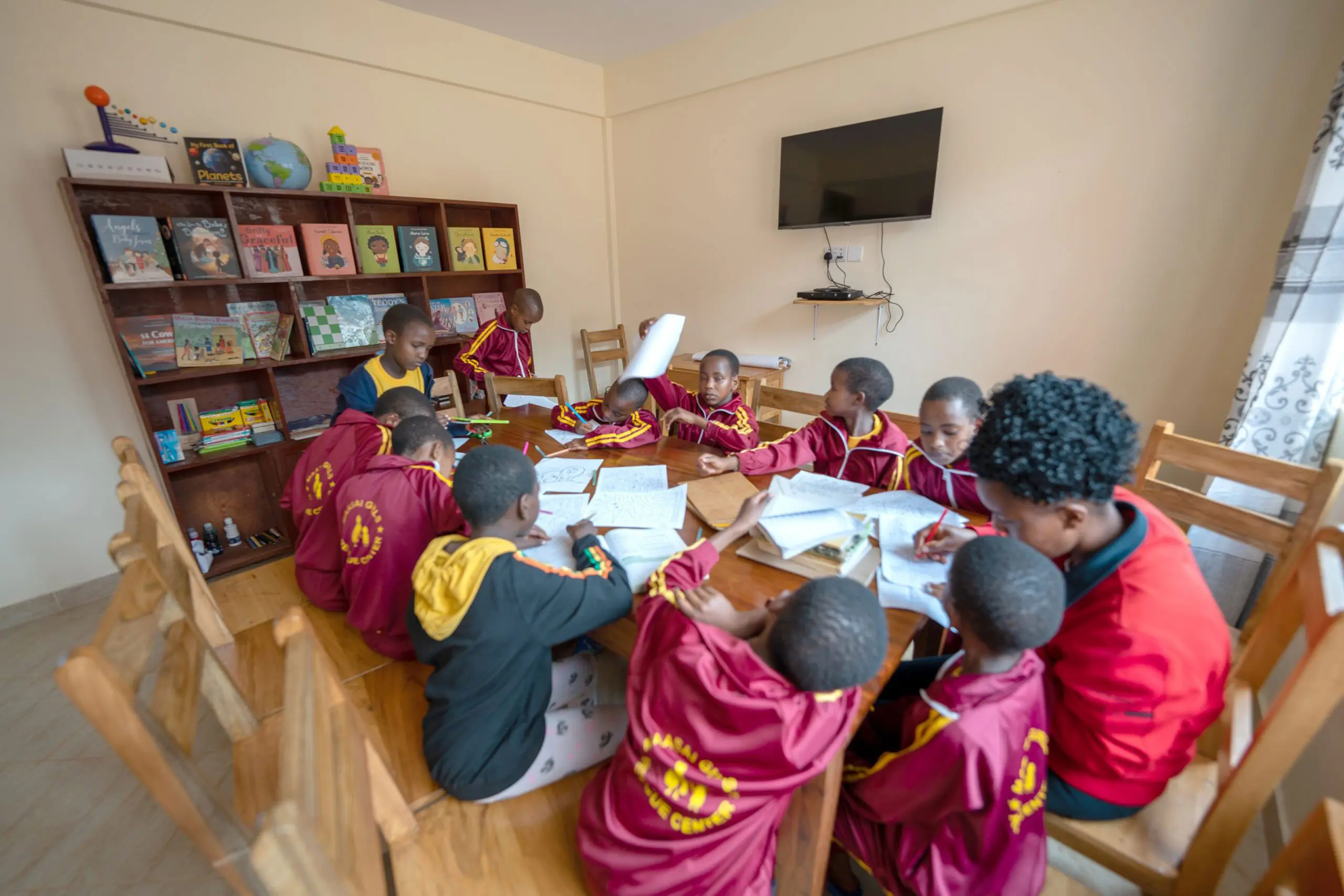 Maasai Girls Rescue Center house mother tutoring young Maasai girls at a long table.