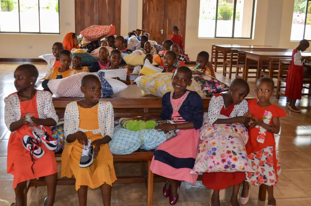 A group of Maasai girls sits on benches indoors, clutching gifts like shoes and pillows. The room, adorned with wooden furniture, is filled with the vibrant colors of their dresses, bringing a festive Christmas charm to the scene.