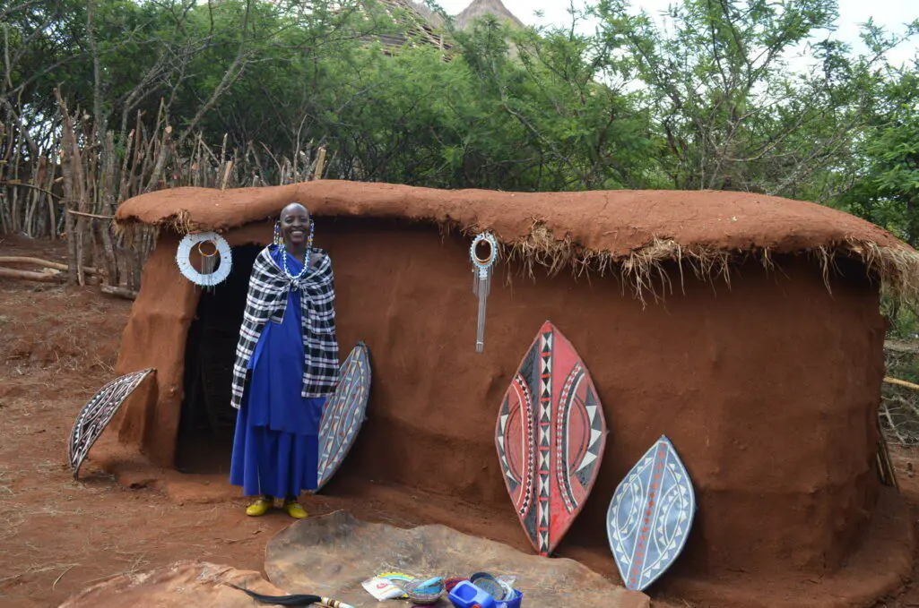 coco, maasai for grandmother, standing in front of a boma decorated with shields and beadwork