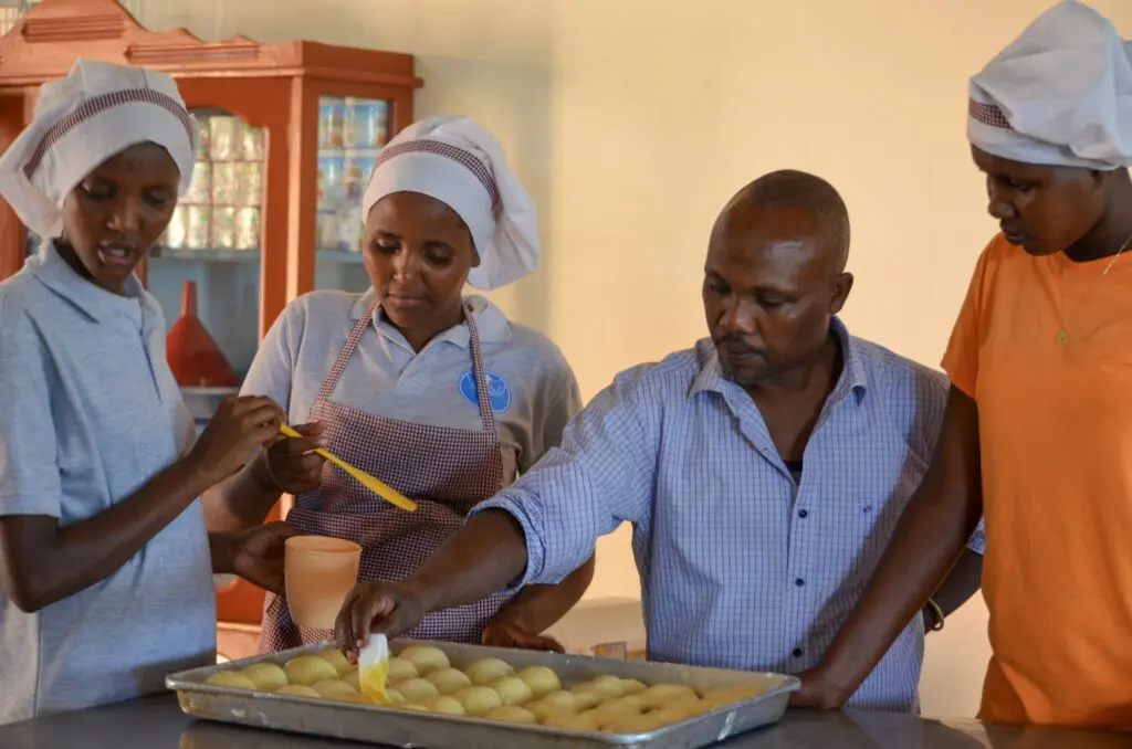 Maasai teen girls learning to bake biscuits, ecolodge chef demostrating buttering technique for biscuit tops 