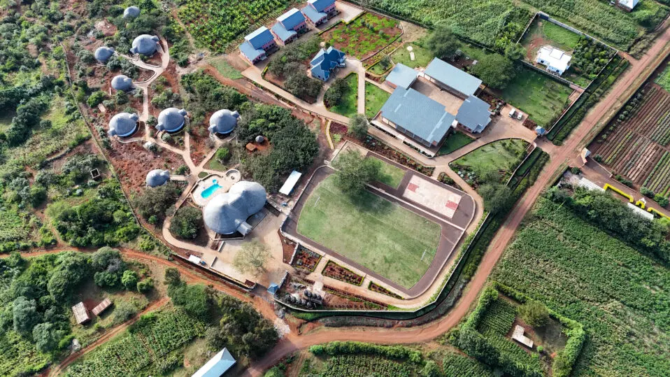 Aerial view of a ecoVillage compound with dome-shaped buildings, rectangular structures, a swimming pool, sports court, and surrounding greenery and farmland.