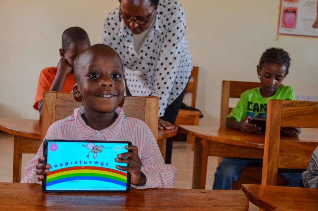 Child smiling with tablet in classroom at Maasai Girls Rescue Center. A new computer teacher recently added to our large family is helping a student in the background.